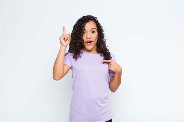 Joven negra sintiéndose orgullosa y sorprendida, apuntando a sí misma con confianza, sintiéndose como la número uno exitosa contra la pared blanca