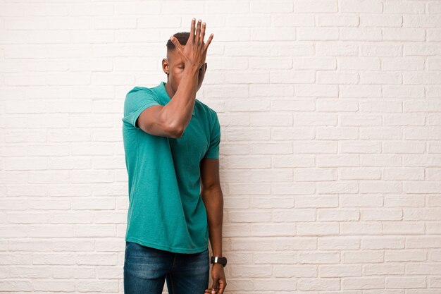 Joven negro afroamericano levantando la palma de la mano a la frente pensando uy, después de cometer un error estúpido o recordar, sentirse tonto en la pared de ladrillo