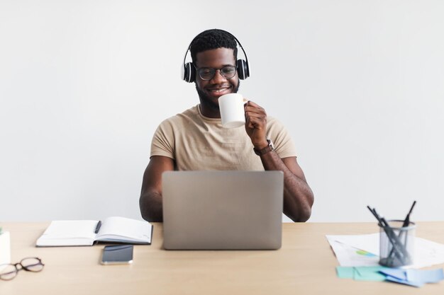 Un joven negro alegre con auriculares bebiendo café charlando en línea en una laptop en el escritorio contra