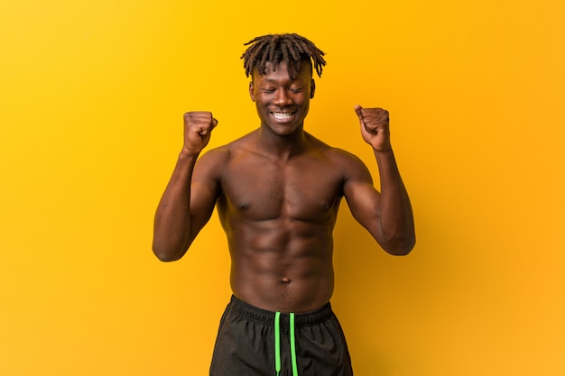 Joven negro sin camisa vistiendo traje de baño levantando el puño, sintiéndose feliz y exitoso. Concepto de victoria