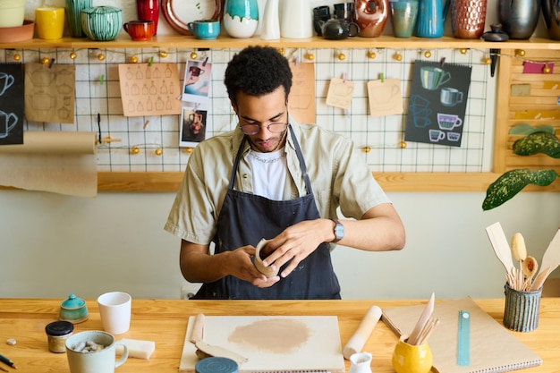 Joven negro creativo en ropa de trabajo creando un artículo de arcilla por mesa en el taller