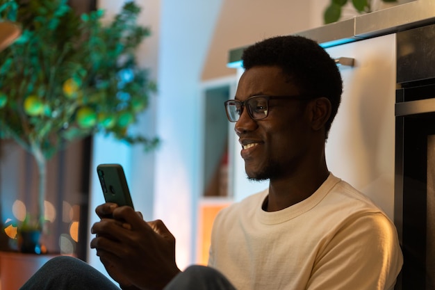 Un joven negro feliz usando su teléfono inteligente mientras se relaja y pasa su tiempo libre en una casa acogedora