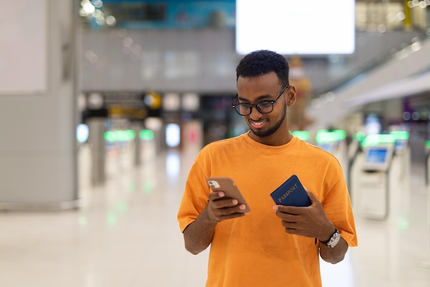 Joven negro listo para viajar en la terminal del aeropuerto esperando el vuelo mientras usa el teléfono