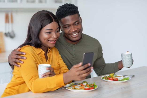 Joven negro y mujer usando un teléfono inteligente mientras disfruta del desayuno en la cocina