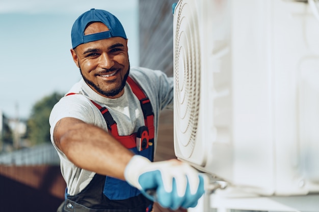 Joven negro reparador comprobando una unidad de aire acondicionado exterior