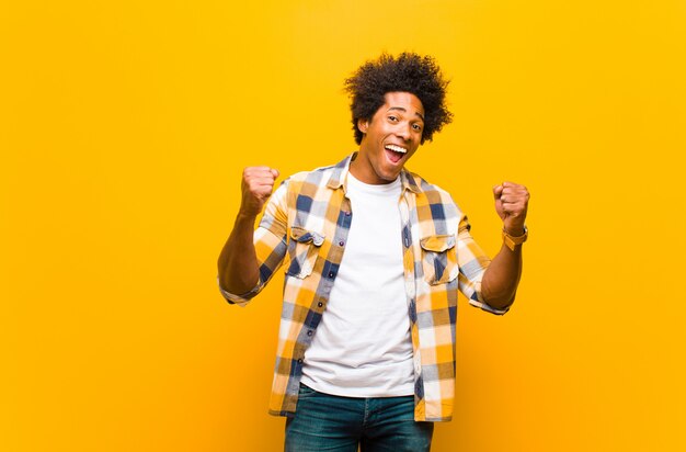 Joven negro sintiéndose sorprendido, emocionado y feliz, riendo y celebrando el éxito, diciendo ¡guau! contra la pared naranja