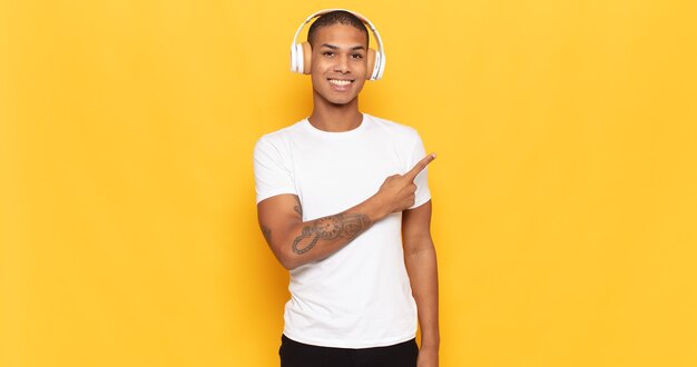 Joven negro sonriendo alegremente, sintiéndose feliz y apuntando hacia un lado y hacia arriba, mostrando el objeto en el espacio de la copia