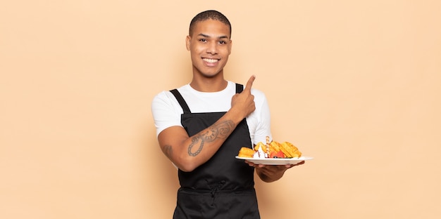 Joven negro sonriendo alegremente, sintiéndose feliz y apuntando hacia un lado y hacia arriba, mostrando el objeto en el espacio de la copia