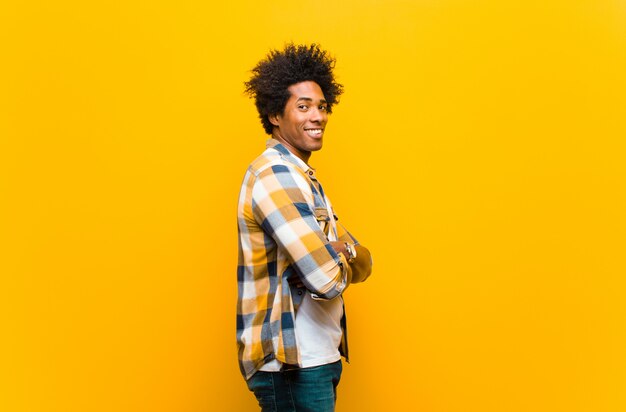 Joven negro sonriendo con los brazos cruzados y una expresión feliz, segura y satisfecha, vista lateral contra la pared naranja