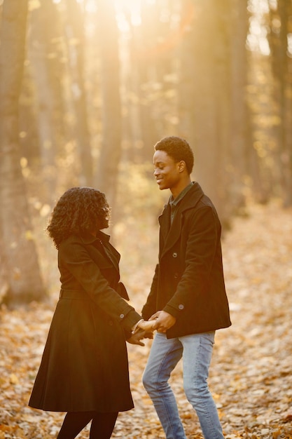 Joven negro y su novia bailando. Pareja romántica caminando en el parque de otoño al atardecer. Hombre y mujer con abrigos negros.