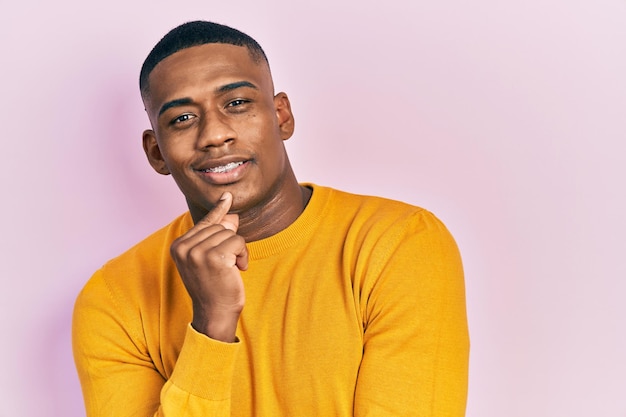 Joven negro con suéter amarillo casual sonriendo mirando confiado a la cámara con los brazos cruzados y la mano en la barbilla. pensando en positivo.