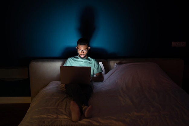 Un joven negro trabaja en una computadora portátil acostado en la cama por la noche en la oscuridad mira atentamente a la pantalla