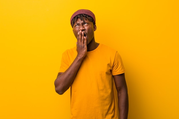 Joven negro vistiendo rastas en amarillo bostezando mostrando un gesto cansado con la boca con la mano.