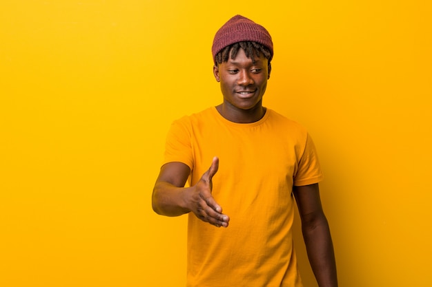 Joven negro vistiendo rastas sobre pared amarilla estirando la mano en gesto de saludo.