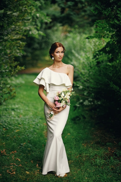 Joven novia hermosa con ramo de flores en un vestido blanco de pie solo al aire libre