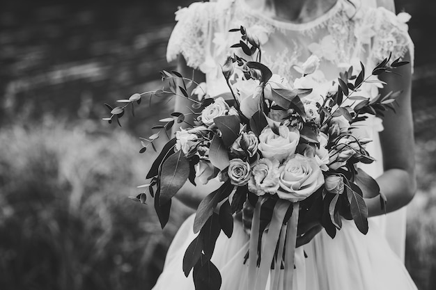 Joven novia hermosa con un vestido elegante está de pie y sosteniendo un ramo de flores y verduras con cinta en la naturaleza en el parque Foto en blanco y negro