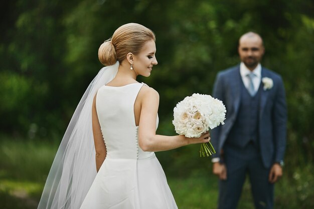 Joven novia de moda en elegante vestido blanco con un ramo de flores en la mano se encuentra al aire libre y esperando a un novio