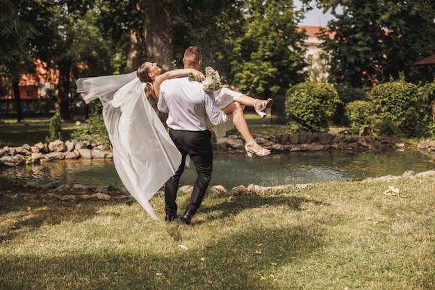 Joven novio alegre llevando y girando a su nueva esposa en la naturaleza el día de la boda.