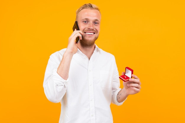 Joven novio con una camisa blanca hace una propuesta de matrimonio a la novia con un anillo en una caja y hablando por teléfono en amarillo