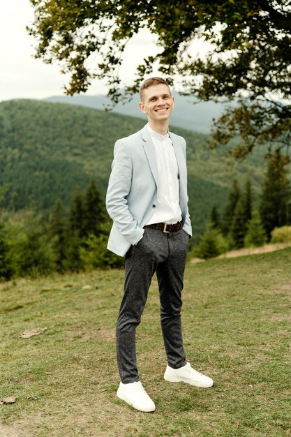 Un joven novio con una chaqueta azul se encuentra cerca de un árbol con el telón de fondo de las montañas