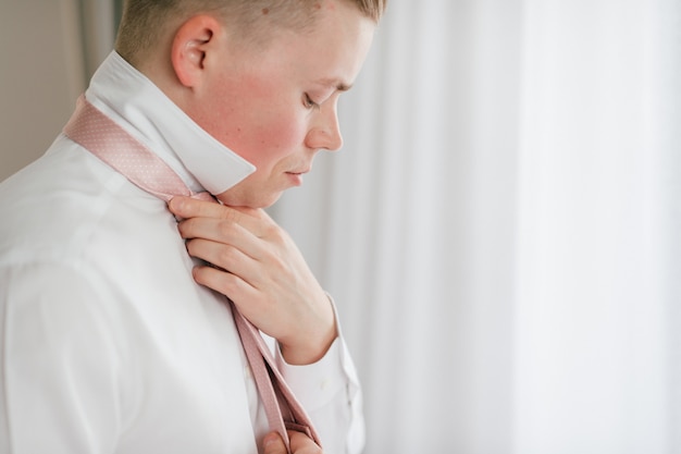 Joven novio preparándose. Prometido por la mañana en casa. Novio arreglando su corbata