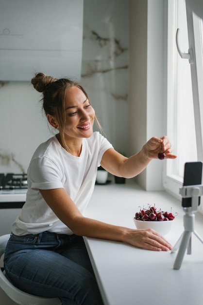 Una joven nutricionista bloguera realiza una consulta o una lección abierta por enlace de video sobre los beneficios de las bayas frescas sentadas en la cocina con un plato de cerezas