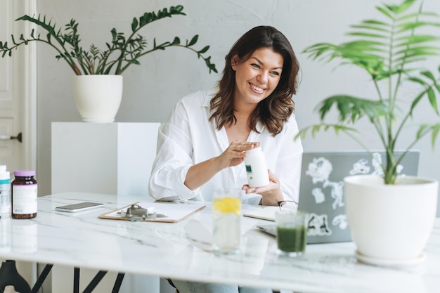 Joven nutricionista morena sonriente de talla grande con camisa blanca que trabaja en una laptop en la mesa con una planta en la oficina moderna y luminosa El doctor se comunica con el paciente en línea