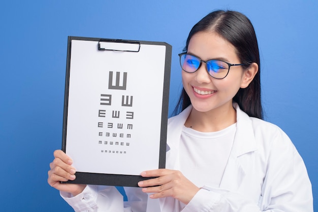 Una joven oftalmóloga con gafas sosteniendo tabla optométrica sobre pared azul