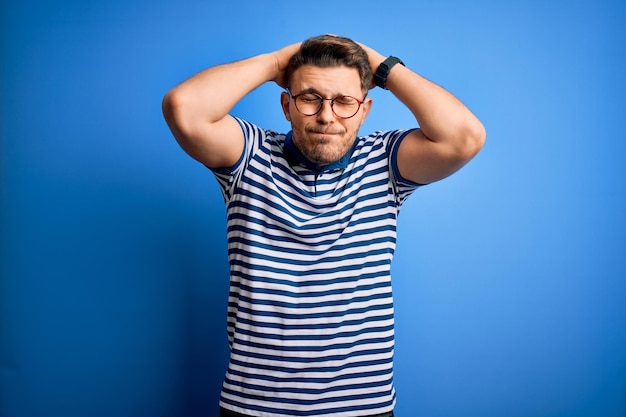 Joven con ojos azules con gafas y camiseta casual a rayas sobre fondo azul que sufre de dolor de cabeza desesperado y estresado por el dolor y la migraña Manos en la cabeza