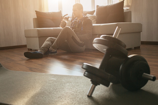 Joven ordinario practicar deporte en casa. El primer año de entrenamiento cansado se sienta solo en el piso y descansa después del ejercicio. Bebe agua y relájate. Par de pesas acostado en la estera. Hombre ordinario después del entrenamiento.