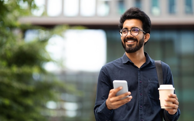 Joven oriental feliz bebiendo café para llevar usando el teléfono al aire libre