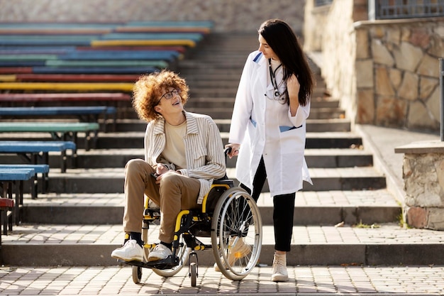 Un joven paciente en un carrito le sonríe a su médico Una enfermera atractiva está hablando con un paciente al aire libre ayudándolo a bajar las escaleras