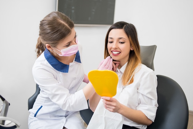 Joven paciente feliz y dentista con sonda están revisando los dientes en el espejo después del procedimiento dental en el consultorio dental