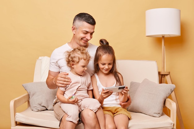 Un joven padre amoroso sentado en el sofá con sus hijas pequeñas lindas viendo videos divertidos en el teléfono inteligente juntos un padre feliz y cariñoso y una niña pequeña usando el teléfono móvil en casa
