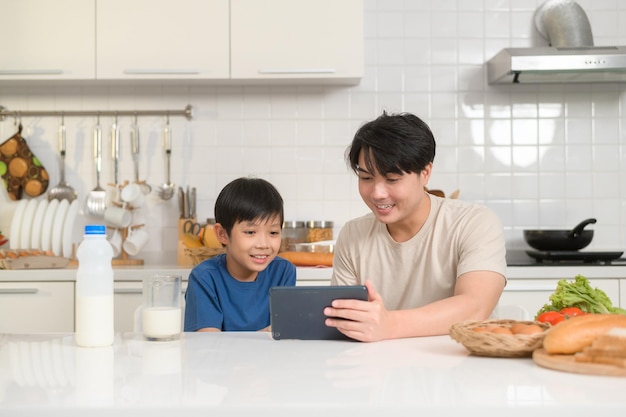 Joven padre asiático y su hijo usando una tableta digital disfrutando juntos en la cocina en casa