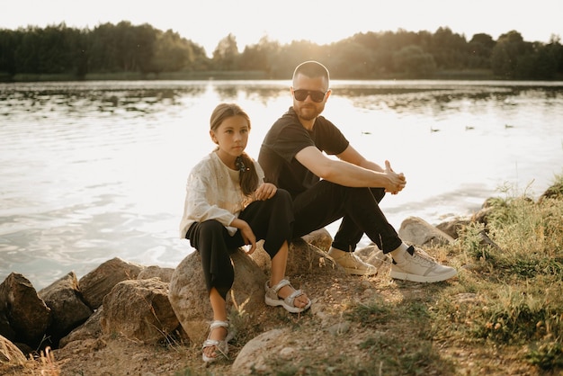 Un joven padre con barba y gafas de sol posa sobre piedras con su linda hija en la costa del lago. Elegante familia monoparental de vacaciones al atardecer.