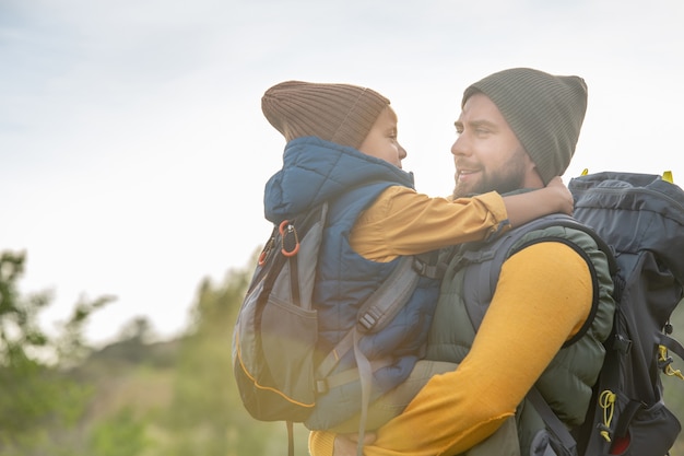 Foto joven padre cariñoso con mochila sosteniendo a su pequeño y lindo hijo en las manos contra el entorno natural contra el cielo, la hierba verde y los arbustos