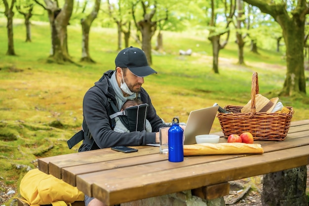 Un joven padre caucásico teletrabajo sentado en una mesa de picnic con computadora en el bosque