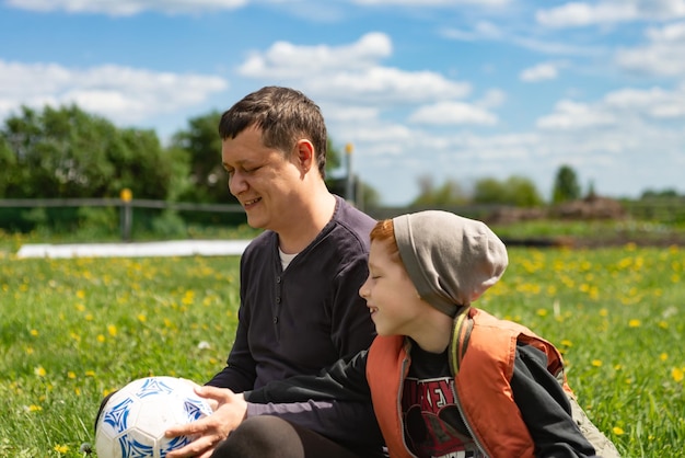 Joven padre e hijo se sientan en el césped en verano después de jugar al fútbol juntos retrato de estilo de vida