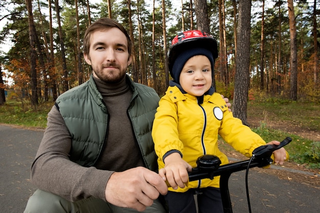 Joven padre feliz en ropa casual en cuclillas por su pequeño y lindo hijo en el casco de seguridad sosteniendo las manijas de la bicicleta de equilibrio mientras se enfría en el parque