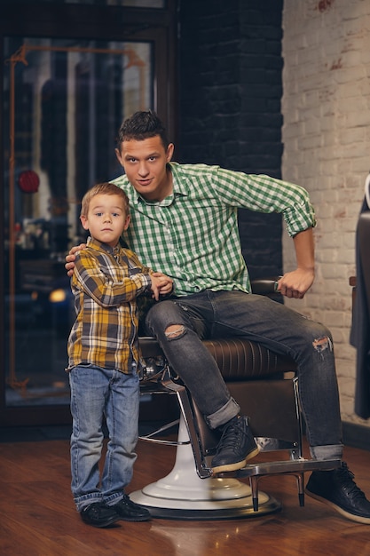 Foto joven padre guapo y su pequeño hijo con estilo en la barbería esperando peluquero
