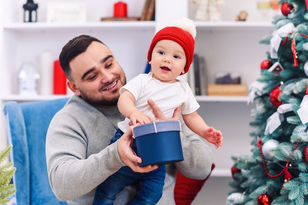 El joven padre se sienta en una silla cómoda con un niño divertido y le da una caja de regalo en la habitación maravillosamente decorada para celebrar la Navidad.