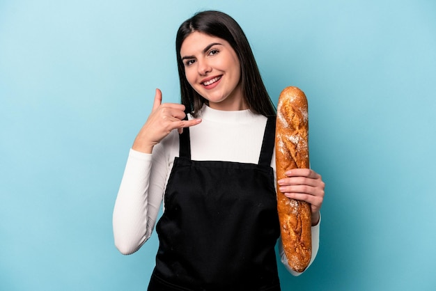 Joven panadero caucásico sosteniendo una hogaza de pan aislado de fondo azul mostrando un gesto de llamada de teléfono móvil con los dedos