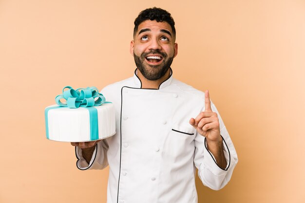 Joven panadero latino sosteniendo un pastel aislado apuntando al revés con la boca abierta.