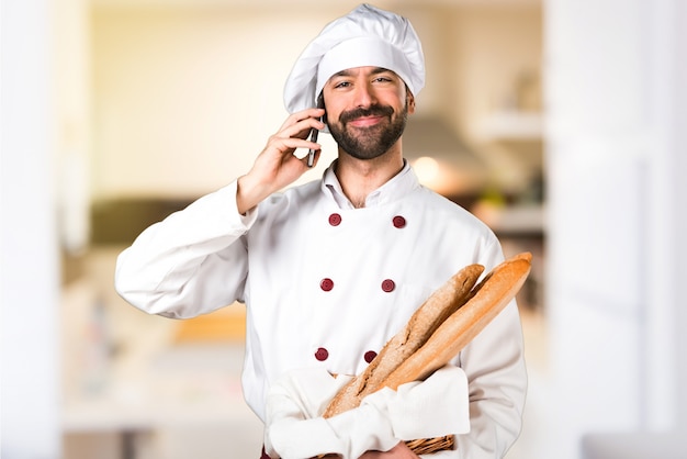 Joven panadero sosteniendo un poco de pan y hablando con el móvil en la cocina