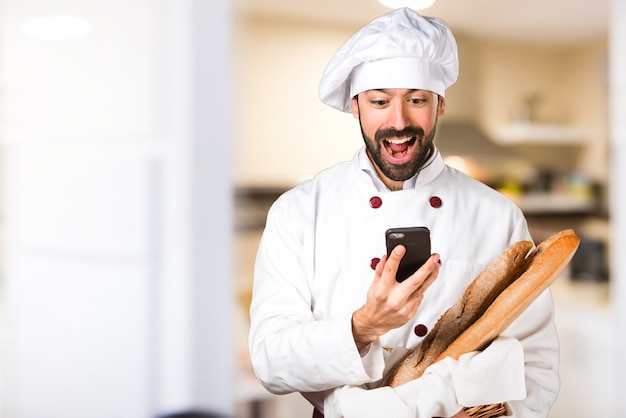 Joven panadero sosteniendo un poco de pan y hablando con el móvil en la cocina