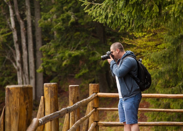 Joven con pantalones azules y zapatillas rojas con mochila negra parado en la veranda de madera y tomando fotos en el fondo del bosque verde