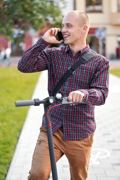 Joven con pantalones casuales y camisa parado al lado de su scooter eléctrico, hablando por teléfono móvil