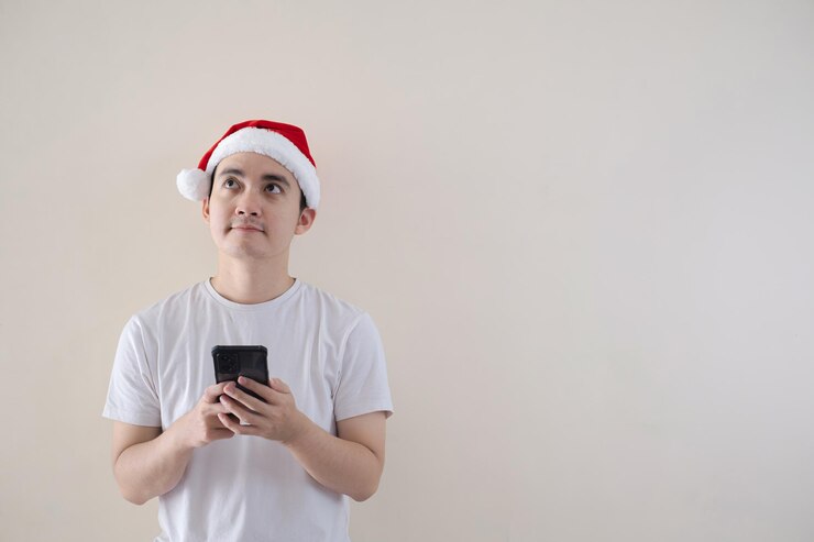 El joven Papá Noel asiático está confundido mientras sostiene un teléfono inteligente y mira hacia arriba