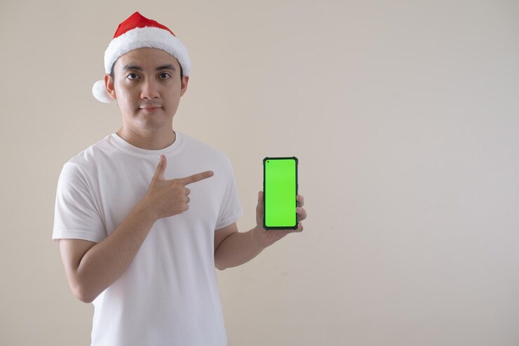El joven Papá Noel asiático está señalando su teléfono inteligente de pantalla verde mientras mira a la cámara